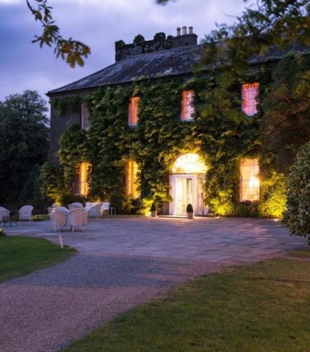 Bij zonsondergang staat Ballymaloe House als een historisch met klimop bedekt landhuis, warm verlicht met gele lichten. Een grindpad leidt naar de uitnodigende witte deur. Op het stenen terras nodigen rieten stoelen en tafels gasten uit om te ontspannen. De bewolkte lucht vormt een aanvulling op de verzorgde gazons, struiken en bomen van het hotel. | Echt Ierland Vakanties